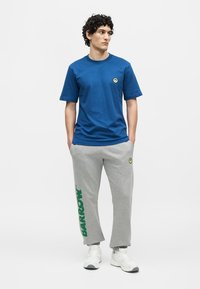 Jeune homme aux cheveux bouclés portant un t-shirt bleu et un pantalon de survêtement gris avec un texte vert et des baskets blanches, debout mains dans les poches.
