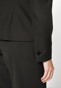 Gros plan de la main d’une personne à côté du poignet d’une veste de costume noire ajustée avec un seul bouton noir.