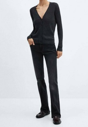 Persoon draagt een getailleerde donkergrijze cardigan met knopen, zwarte jeans met rechte pijpen en donkere loafers, staand met één hand in de zak.