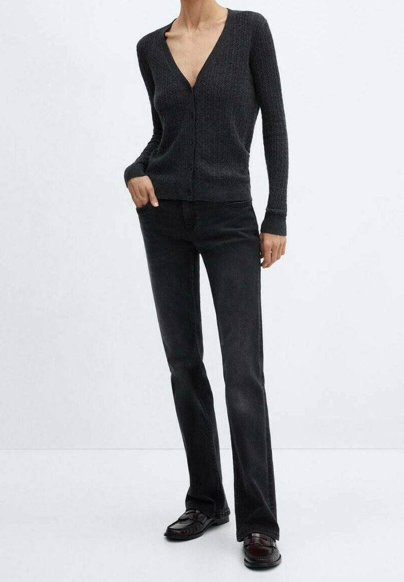 Personne portant un cardigan gris foncé ajusté boutonné, un jean noir droit et des mocassins foncés, debout avec une main dans la poche.