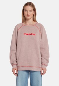 Sweatshirt en polaire rose avec des coutures rouges, portant le mot "mankind" en lettres rouges en gras sur la poitrine. Coupe décontractée.