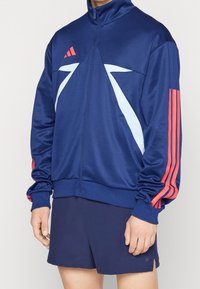 Man wearing navy blue track jacket with light blue geometric patterns and red stripes on sleeves, paired with navy shorts.