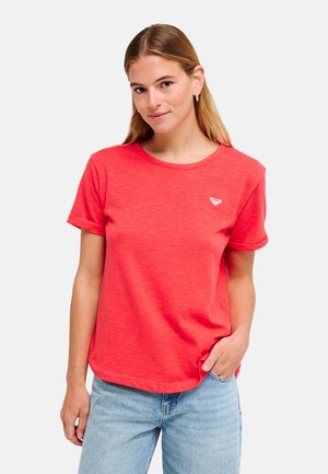 Kobieta o jasnobrązowych włosach, ubrana w czerwoną koszulkę z krótkim rękawem i jasnoniebieskie dżinsy, stojąca z jedną ręką w kieszeni.