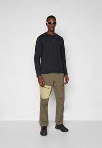 Zwarte langemouwen shirt met logo, khaki cargobroek met een beige ritszak, en zwarte schoenen, aangevuld met lichtgekleurde zonnebril.