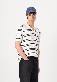 Kremstripet polo-skjorte med korte ermer, med svarte horisontale striper og V-hals, kombinert med mørke jeans og en blå caps.
