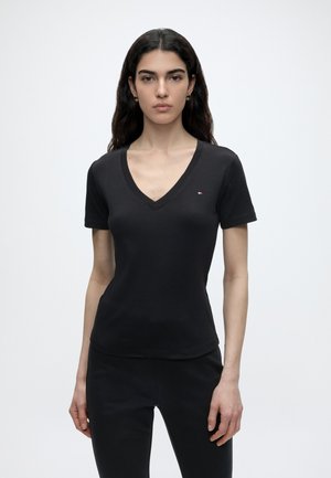 Kobieta o długich ciemnych włosach, ubrana w czarną koszulkę z krótkim rękawem i dekoltem w serek oraz czarne spodnie, stojąca na tle jednolitego jasnego tła.