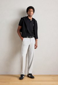 Zwarte poloshirt met logo op de borst, grijze broek, zwarte riem en zwarte sneakers met witte strepen. Model staat tegen een neutrale achtergrond.