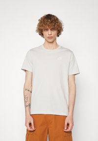 Béžové bavlnené tričko s krátkymi rukávmi, okrúhlym výstrihom a bielym logom Nike na hrudi. Voľný strih, hladká textúra a minimalistický dizajn.