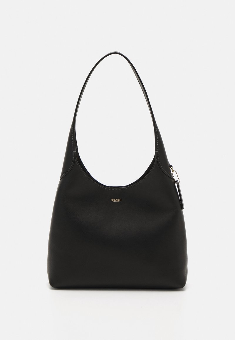 Sac à bandoulière en cuir noir avec une seule bandoulière courbée, un petit logo doré Coach et une étiquette métallique suspendue à la poignée, sur un fond uni.
