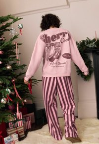 Różowa bluza z retro grafiką wiśni na plecach oraz różowymi i ciemnoczerwonymi spodniami od piżamy w paski. Na tle choinki bożonarodzeniowej.