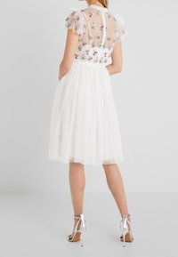 Valkoinen mekko, jossa on läpinäkyvä, kirjailtu yläosa kukkakuvioilla ja röyhelöiset lyhyet hihat; kerroksellinen tyllihame; yhdistetty hopeisiin korkokenkiin.