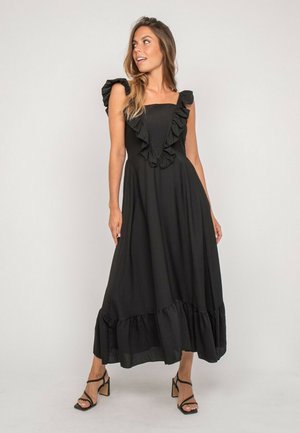 Femme portant une robe maxi noire sans manches avec des volants au niveau de l'encolure et de l'ourlet, associée à des sandales noires à talons blocs et brides.