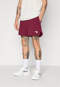 Pantalones cortos deportivos marrón con un logo blanco, con bolsillos a los lados, llevados con zapatillas blancas y calcetines hasta los tobillos. Presenta tatuajes visibles en las piernas.