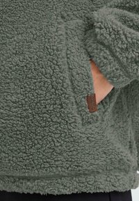 Flauschige grüne Fleecejacke mit einer Tasche und einem braunen Lederschild. Der Stoff hat eine weiche, strukturierte Oberfläche für wärme.