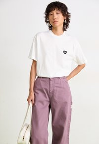 T-shirt en coton blanc avec un logo noir en forme de cœur sur la poitrine gauche, associé à un pantalon lilas ample et un sac à main blanc.