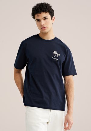 Mladý muž s kučeravými vlasmi nosí tmavomodré tričko s bielym minimalistickým motívom palmy, v kombinácii s bielymi nohavicami, neutrálne pozadie.