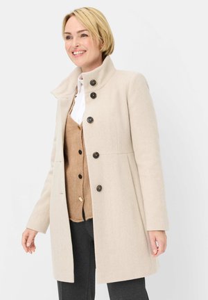 Smiling woman wearing a beige buttoned coat over a tan cardigan and white shirt, standing against a plain light background.