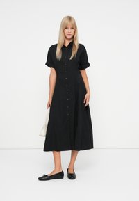 Donna con capelli biondi lisci che indossa un vestito midi nero con bottoni, ballerine nere e tiene in mano una piccola borsa bianca.