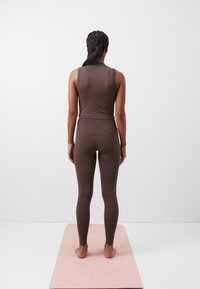 Person med flätat hår som står barfota på en rosa yogamatta, iförd en brun ärmlös topp och matchande leggings, vänd mot en vit vägg.