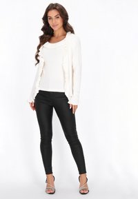 Fehér bordás kardigán fodros részletekkel, egy fehér felsőre rétegezve, fekete slim fit nadrággal és fekete pántos magassarkú cipővel párosítva.