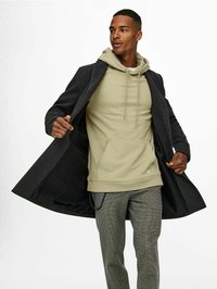 Grijze overjas, olijfgroene hoodie en gemusterde grijze broek. De jas heeft een zachte textuur met een zwarte binnenvoering. Casual gelaagd outfit.