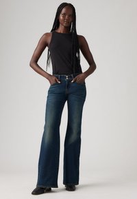 Nainen, jolla on pitkät letit, pukeutuneena mustaan hihattomaan paitoihin, tummansiniset leveälahkeiset farkut ja mustat kengät, seisoo kädet taskuissa.