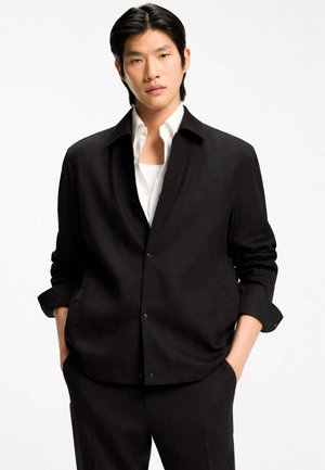 Giovane uomo con capelli neri che indossa una giacca nera abbottonata sopra una camicia bianca, in piedi con le mani nelle tasche contro uno sfondo bianco.