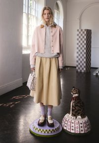 Modella che indossa una giacca corta a righe rosa, felpa grigia con cappuccio e gonna gialla. Scarpe marroni con cinturini bianchi. Tiene una borsa floreale.