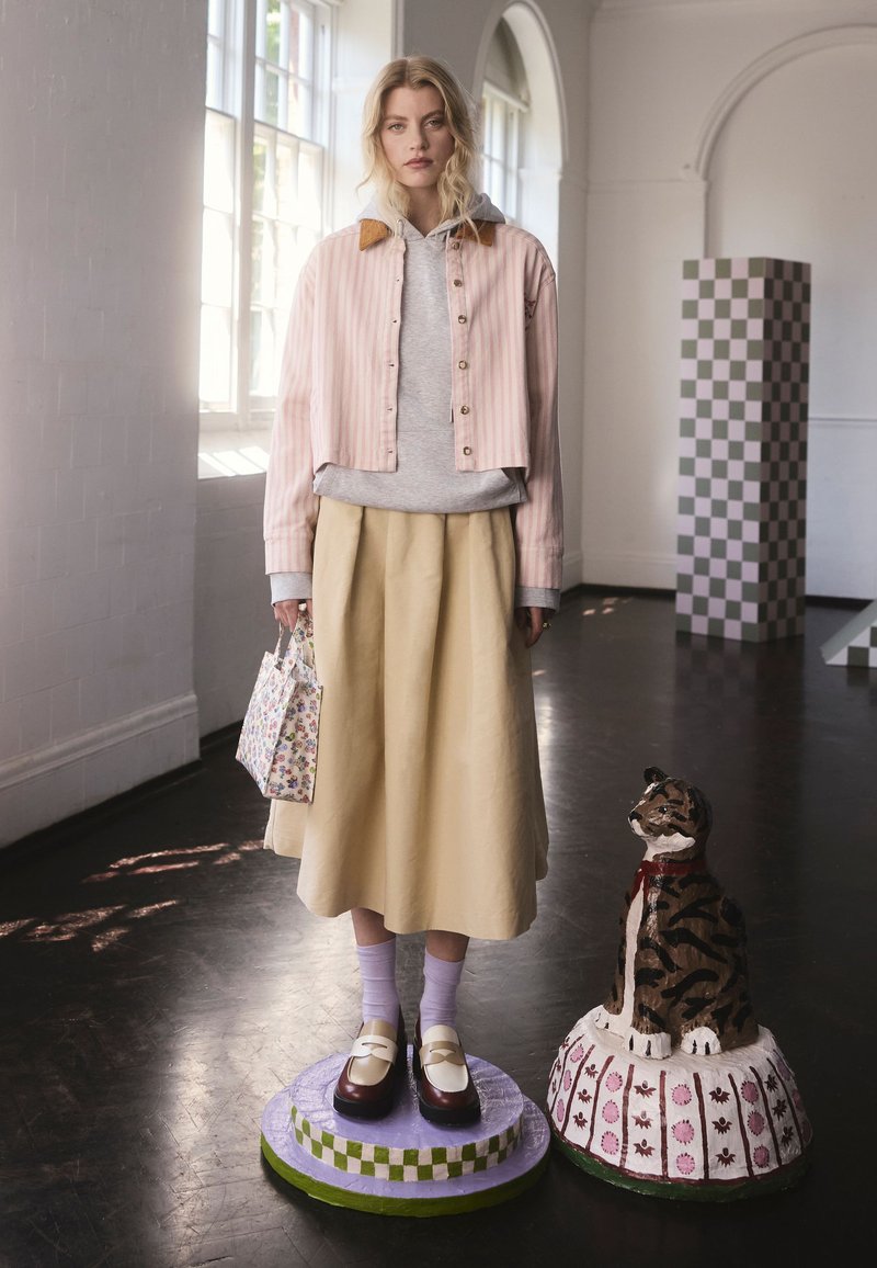 Modella che indossa una giacca corta a righe rosa, felpa grigia con cappuccio e gonna gialla. Scarpe marroni con cinturini bianchi. Tiene una borsa floreale.