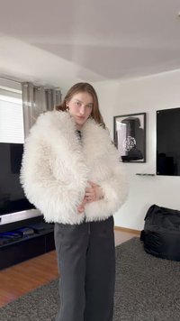Hvit jakke i imitert pels med fluffy tekstur, bred krage og senkede skuldre, kombinert med mørke, løstsittende bukser.