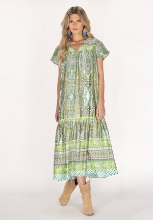 Langes, locker sitzendes Kleid in Grün und Blau mit komplizierten Mustern. Kurze Ärmel und eine Gathernaht am Saum. Getragen mit beigen Stiefeln.