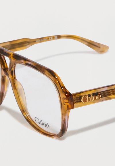 Rudos, permatomos kvadratinės saulės akiniai su storomis rėmelėmis ir skaidriais lęšiais. Ant šoninių dalių yra auksinis prekės ženklo logotipas.