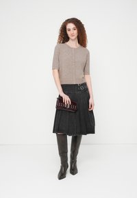 Νεαρή γυναίκα με σγουρά μαλλιά, που φοράει μπεζ πουκάμισο με κουμπιά, μαύρη πλισέ φούστα με αγκράφες, μπότες μέχρι το γόνατο και κρατάει σκούρο τσαντάκι χειρός.