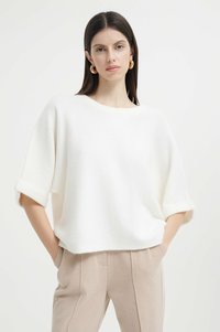 Maglione bianco, ampio e comodo, realizzato in materiale morbido e texturizzato, con maniche corte arrotolate, abbinato a pantaloni beigi. Design semplice e pulito.