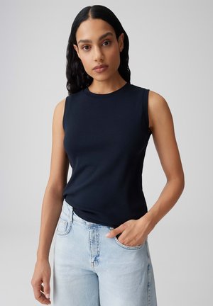 Femme aux longs cheveux noirs portant un haut noir sans manches et un jean bleu clair, debout devant un fond gris uni.