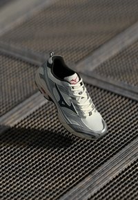 White and gray athletic shoe with black logo resting on metal grating surface with shadows.
