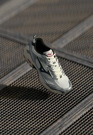 Baskets de sport blanches et grises avec un logo noir reposant sur une surface en grille métallique avec des ombres.