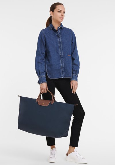 Navy blue nylon tote bag with brown leather accents and handles. Model wears a denim shirt, black pants, and white sneakers.