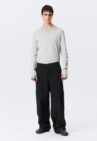 Jeune homme debout, face à l'avant, portant une chemise à manches longues gris clair, un pantalon cargo noir ample et des chaussures noires texturées sur fond blanc.