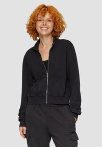 Czarna bluza zapinana na zamek z ribbowymi mankietami i paskiem w talii, z przednią kieszenią i wysokim kołnierzem, wykonana z miękkiego materiału.