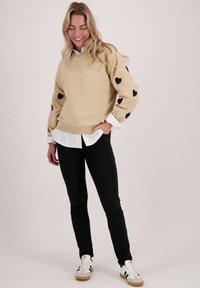 Beige sweatshirt met zwarte hartpatronen op de mouwen, gedragen over een wit shirt, gecombineerd met zwarte broek en casual sneakers.