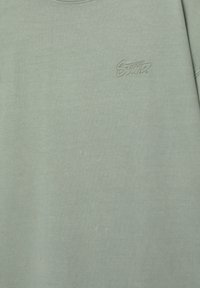 Tricou din bumbac verde deschis cu guler rotund, cu un broderie subtilă. Include un mic logo brodat al brandului pe piept.