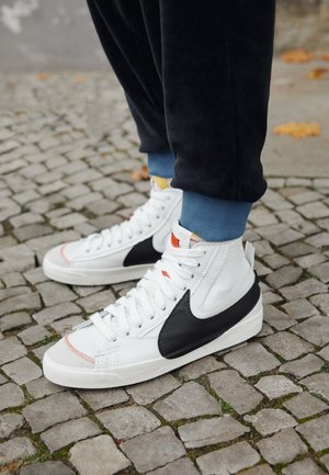 Osoba nosząca białe sneakersy za kostkę z czarnymi logo Swoosh, stojąca na brukowanym chodniku w czarnych spodniach i niebieskich skarpetkach.