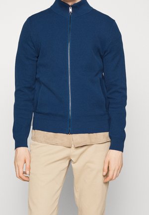 Navy blue zip-up sweater with a high collar, ribbed cuffs, and hem. Woven texture. Paired with light beige pants.