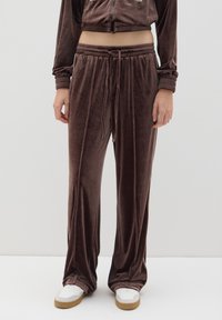 Pantalones de chándal de terciopelo marrón con cintura ajustable, con franjas laterales y un corte relajado. Combinados con zapatillas blancas sobre un fondo liso.