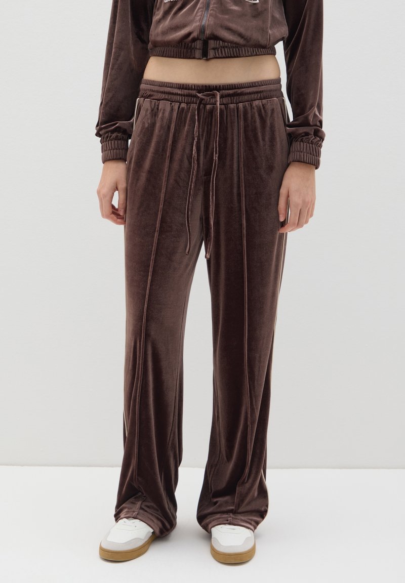 Pantalones de chándal de terciopelo marrón con cintura ajustable, con franjas laterales y un corte relajado. Combinados con zapatillas blancas sobre un fondo liso.