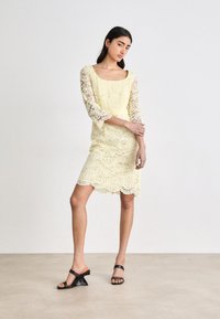 Robe en dentelle jaune clair avec des manches trois-quarts et un ourlet à volants. Portée avec des sandales à talons noires. Texture lisse avec un motif floral.