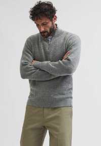 Maglione grigio a maglia con colletto a zip fino al quarto, maniche lunghe e una texture a coste, abbinato a pantaloni beige chiaro.