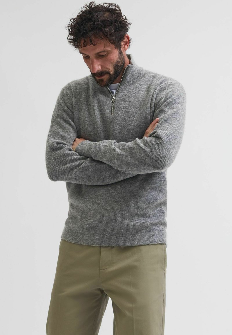 Maglione grigio a maglia con colletto a zip fino al quarto, maniche lunghe e una texture a coste, abbinato a pantaloni beige chiaro.