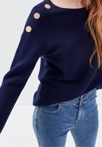 Námořnicky modrý žebrovaný svetr se třemi texturovanými zlatými knoflíky na rameni, doplněný o modré džíny s vysokým pasem, které zdůrazňují pohodlný střih.